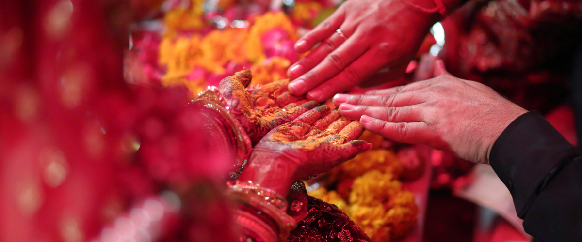 Close-up of two hands joined in a ceremonial gesture, decorated with orange turmeric powder, surrounded by bright red and yellow floral arrangements.