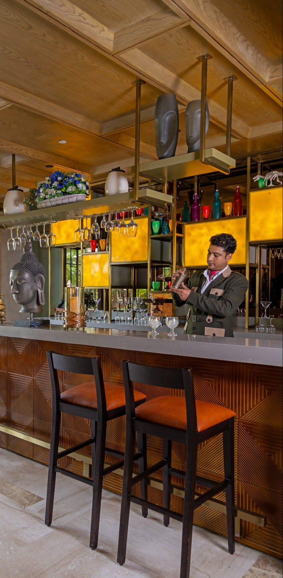 A man in a dark suit stands behind a modern bar counter with three black barstools featuring orange-brown cushions. The bar has a dark wood base, a gray countertop, and backlit yellow-orange cabinets.