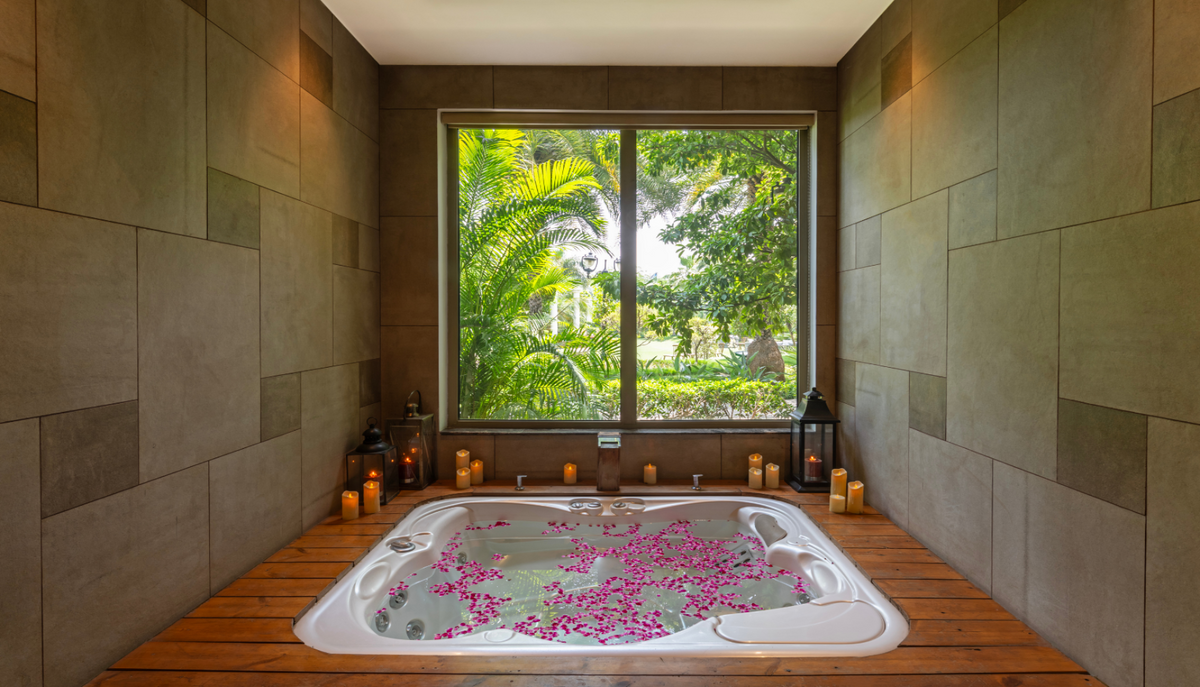 A white hot tub filled with pink flower petals sits on a wooden deck in a spa-like bathroom with stone tile walls and a large window overlooking lush green tropical foliage.